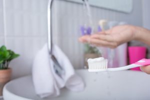 Toothbrush with toothpaste held near a bathroom sink while rinsing hands under running water.